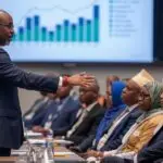 an african conference speaker standing showing economic slides on screen in front of the niger republic government body. mixed dress types suits, boubou, hijab women etc.