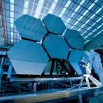 Engineers in protective suits work on telescopic mirrors in a high-tech lab.