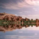 Picturesque reflection of Ouarzazate's historic ksar by the river under a vibrant sky.
