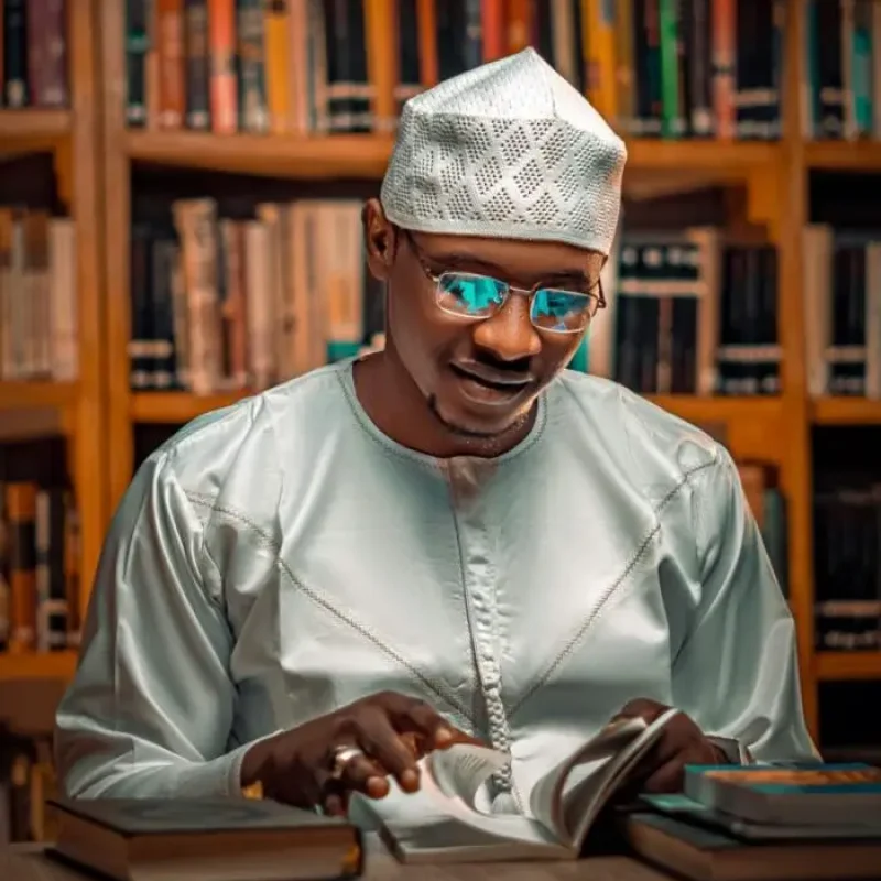 A man in traditional attire reads a book in a library, reflecting focus and style.